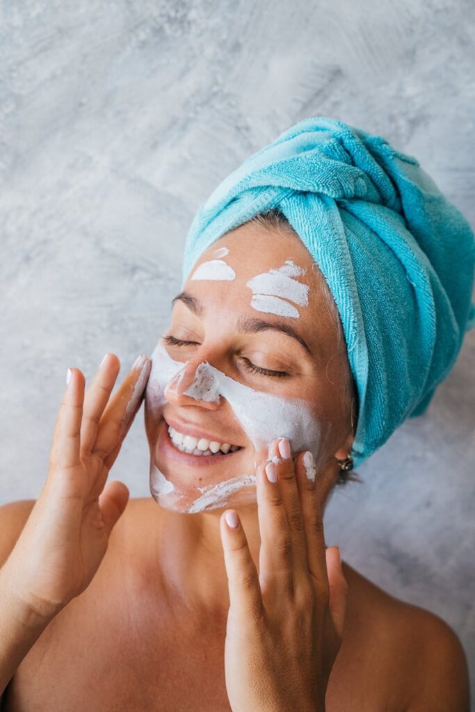 Photo by Anna Tarazevich Smiling woman applying facial cream with eyes closed and towel wrapped around her head.