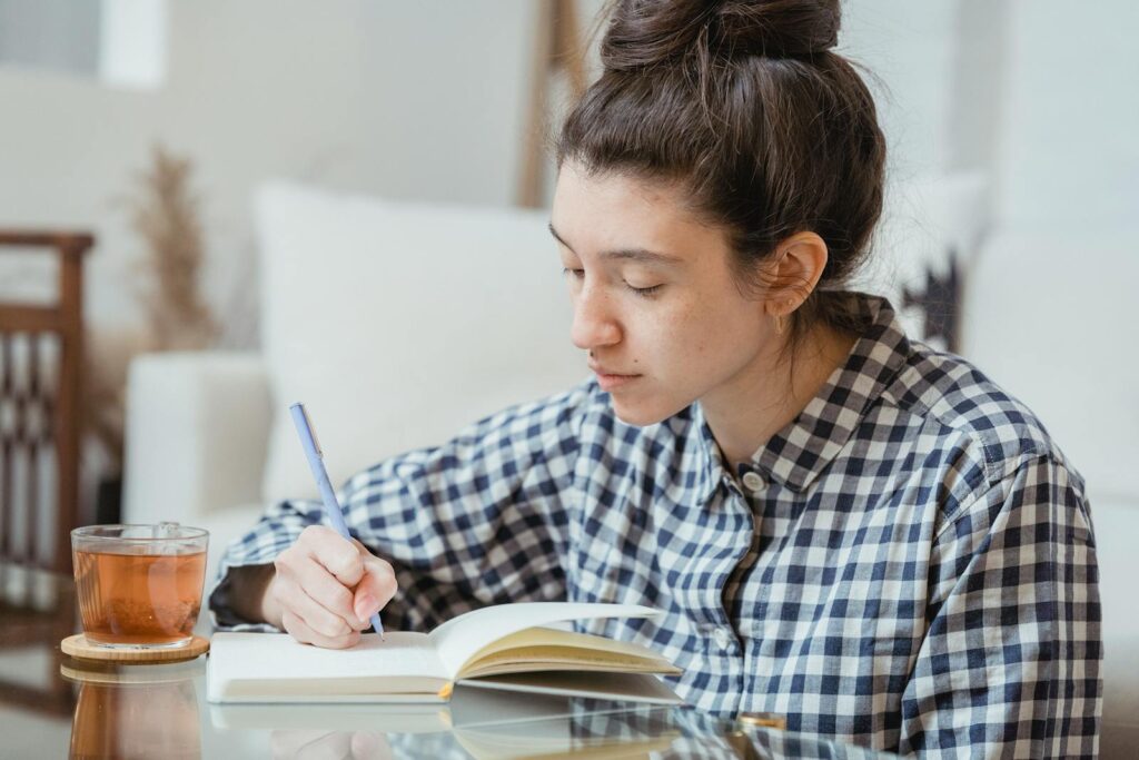 Photo by Miriam Alonso A young woman studying and writing in a notebook with a cup of tea nearby.